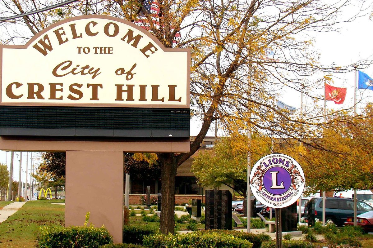 The Welcome to the City of Crest Hill sign in front of City Hall, framed by autumn trees, flags and a Lions International sign