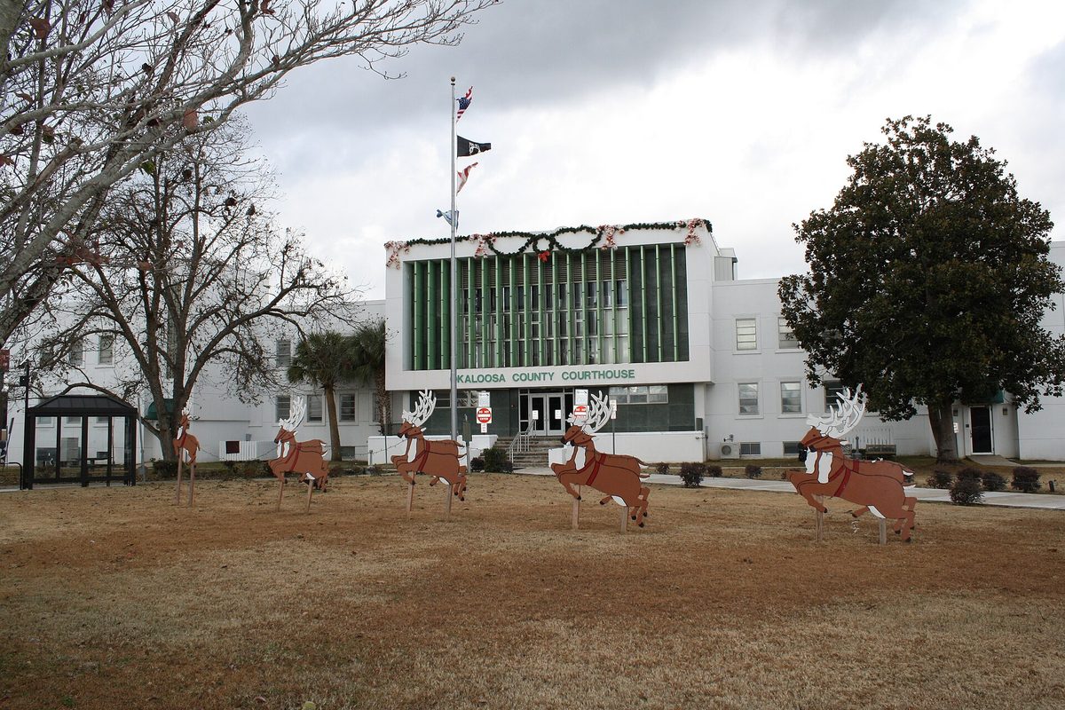 The Okaloosa County Courthouse in Crestview, a white building with a green glass front hung with holiday garland, and wooden reindeer cutouts on the brown lawn