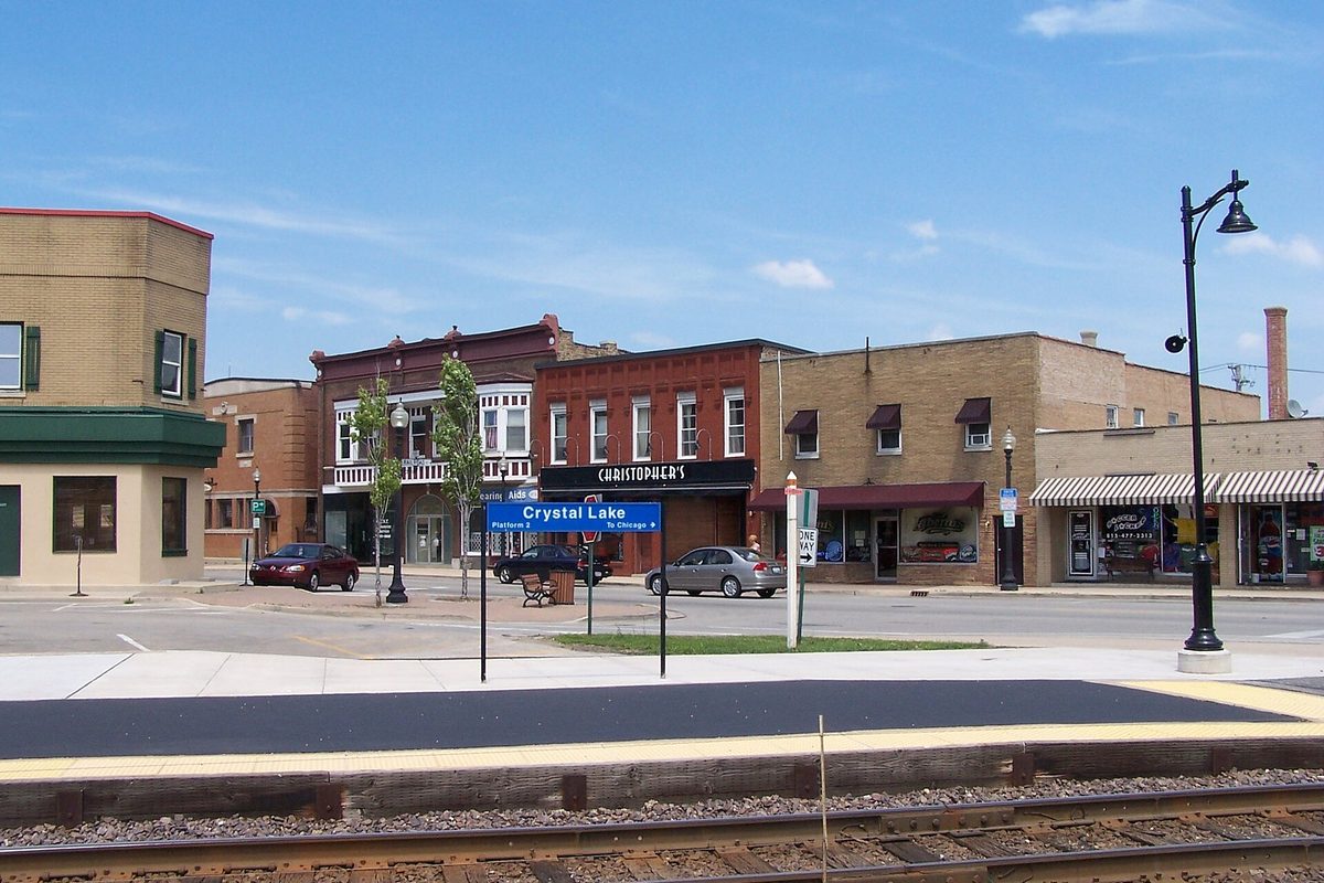 Brick storefronts on Main Street in Crystal Lake seen across the Metra tracks, with a blue Crystal Lake station sign and parked cars