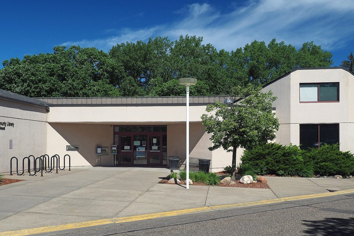 The low beige Rockford Road Library in Crystal, with a glass entrance, a lamp post, a bike rack and tall green trees behind it under a blue sky