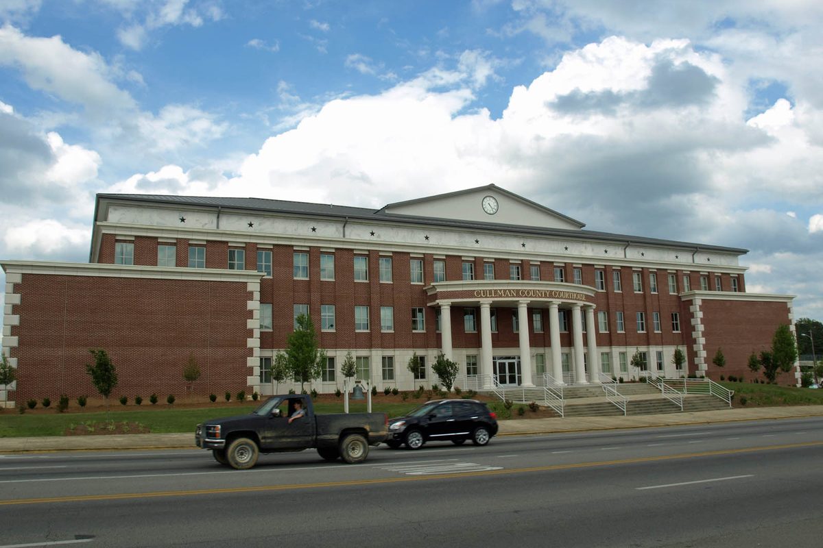 The red brick Cullman County Courthouse with white columns and a clock in its pediment, a pickup and a black sedan passing on the road out front