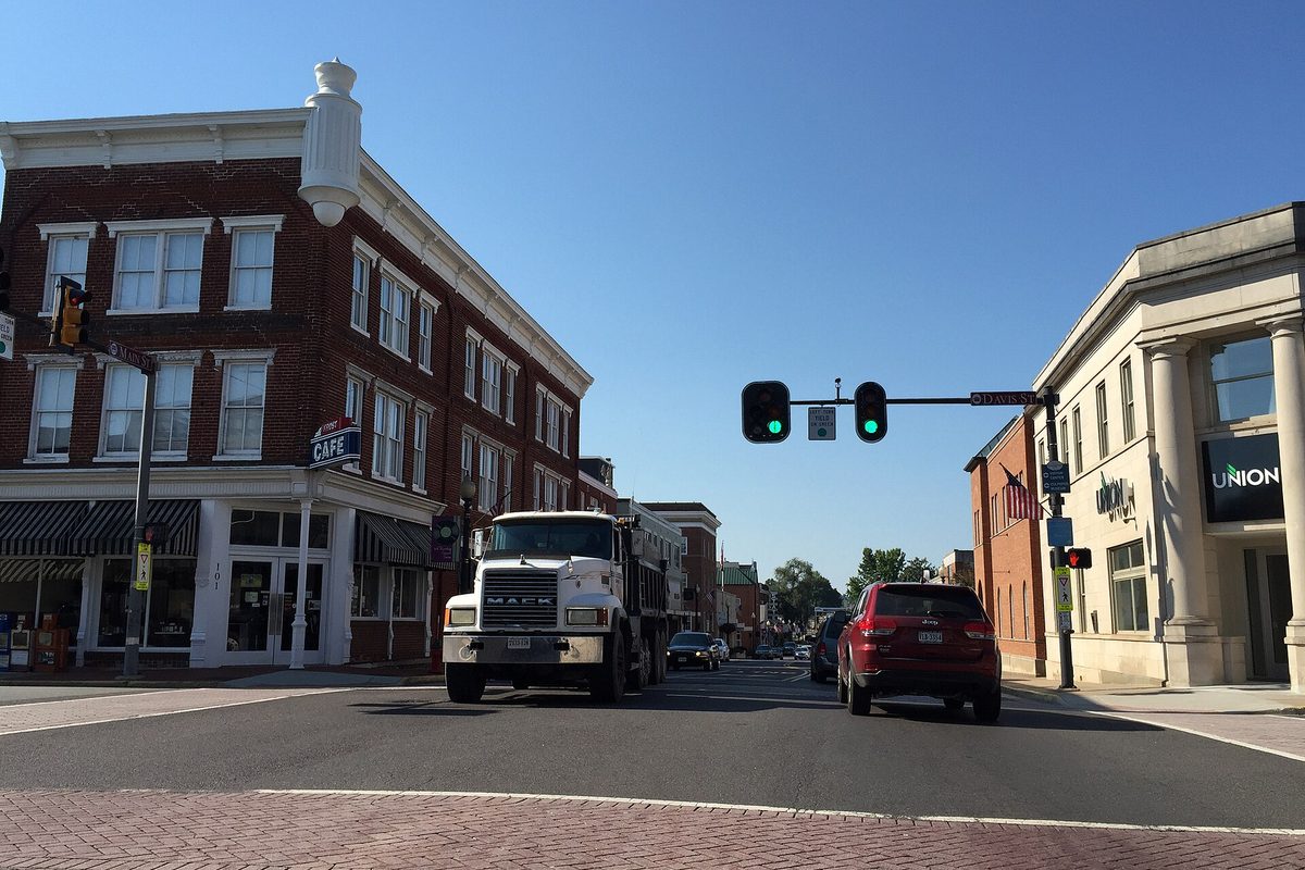Main Street in downtown Culpeper at Davis Street, with a white dump truck and a red SUV at a green light between an old brick building with a cafe sign and a pale stone bank