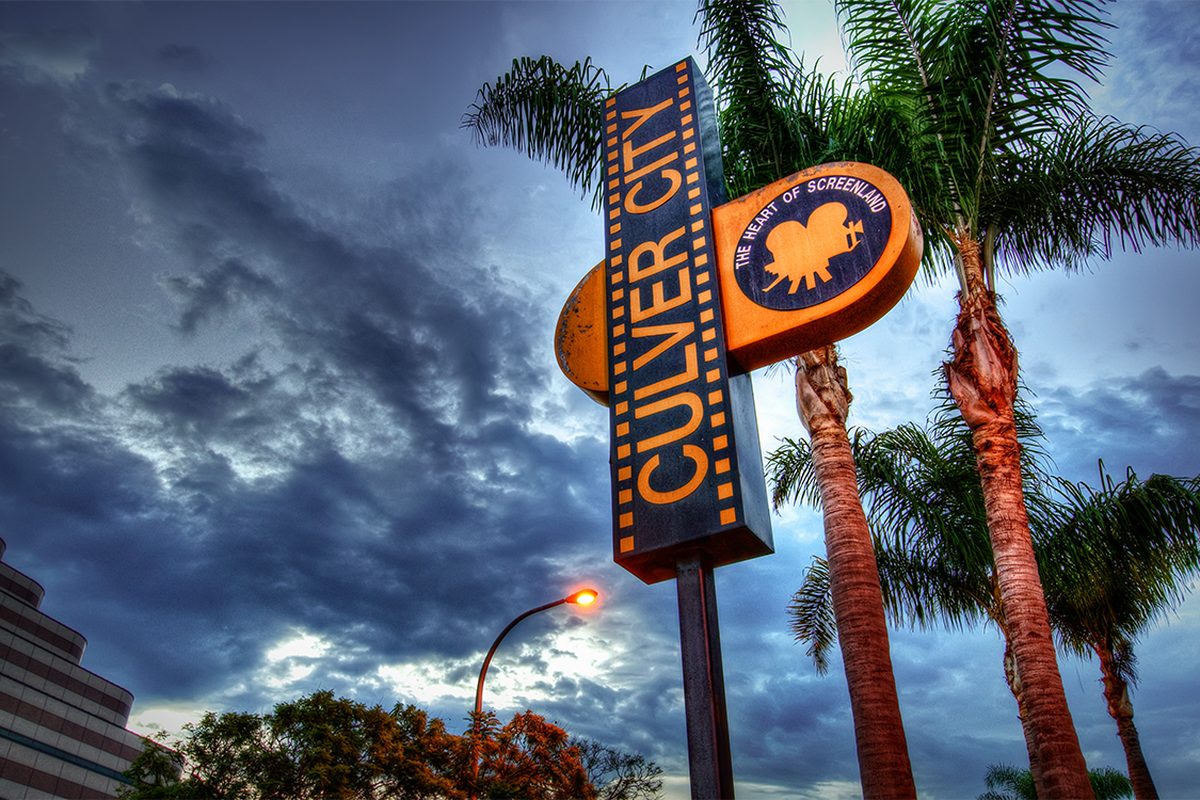 The Culver City sign, The Heart of Screenland, lit at dusk between tall palm trees under a stormy sky