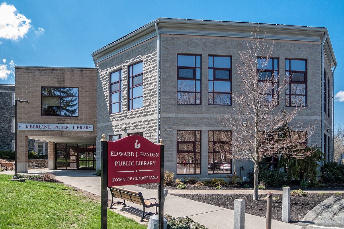 The stone Edward J. Hayden Public Library in Cumberland, with a maroon Town of Cumberland sign and a bench in front and a bare tree beside the entrance