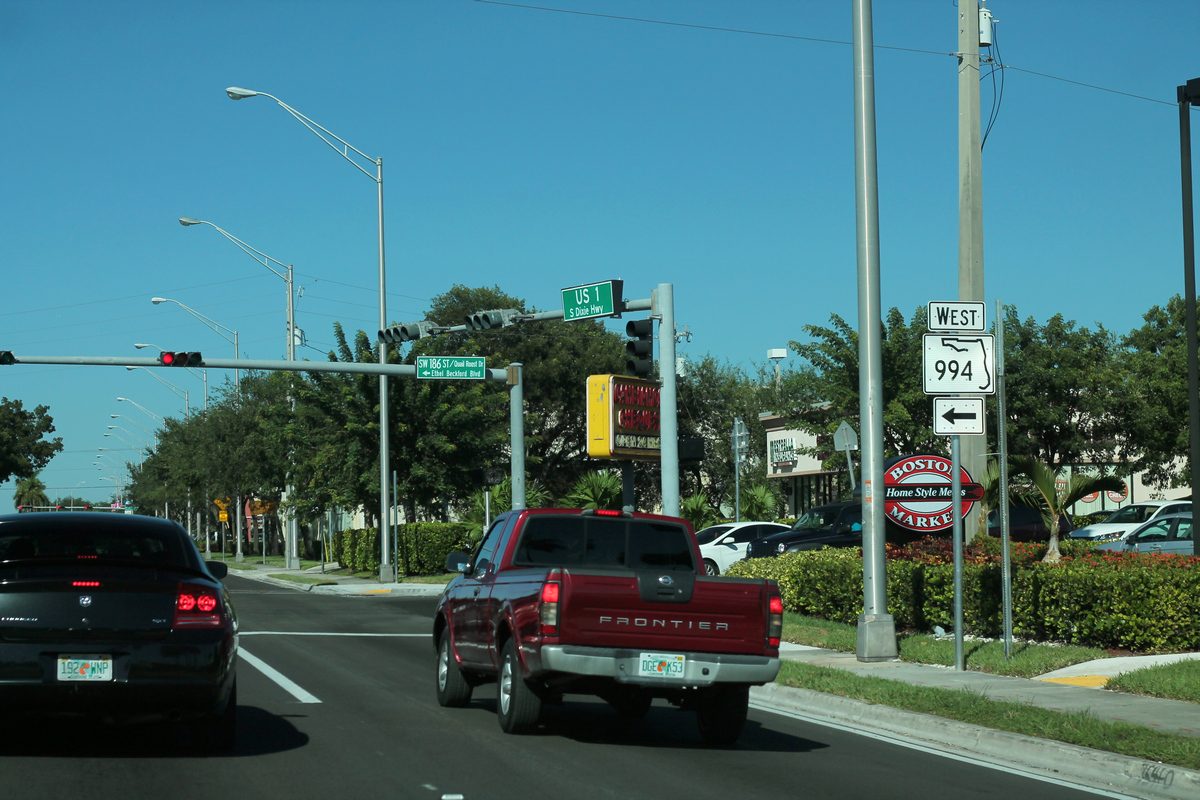 Northbound traffic on US 1 at the sign for State Road 994 West, with a red pickup stopped at the light and palms and shade trees along the road