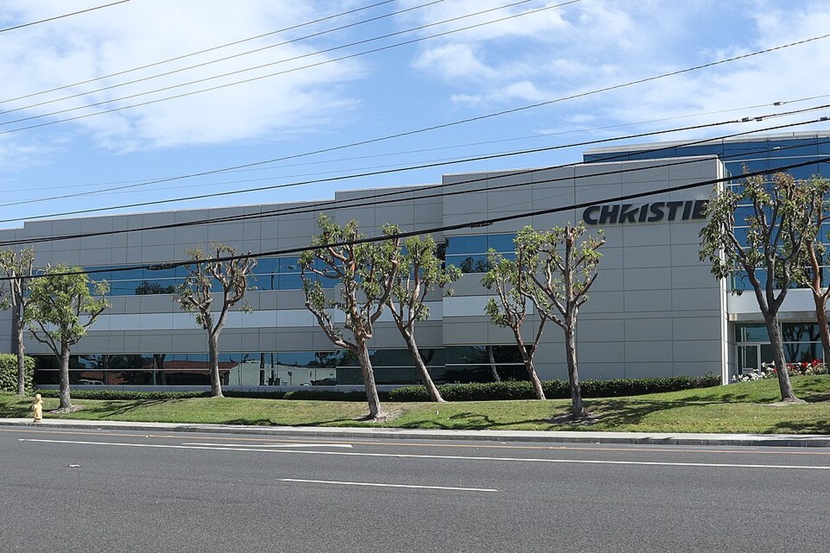 A long gray and glass office building with a Christie sign in Cypress, behind a lawn and a row of pruned trees along a wide, empty street