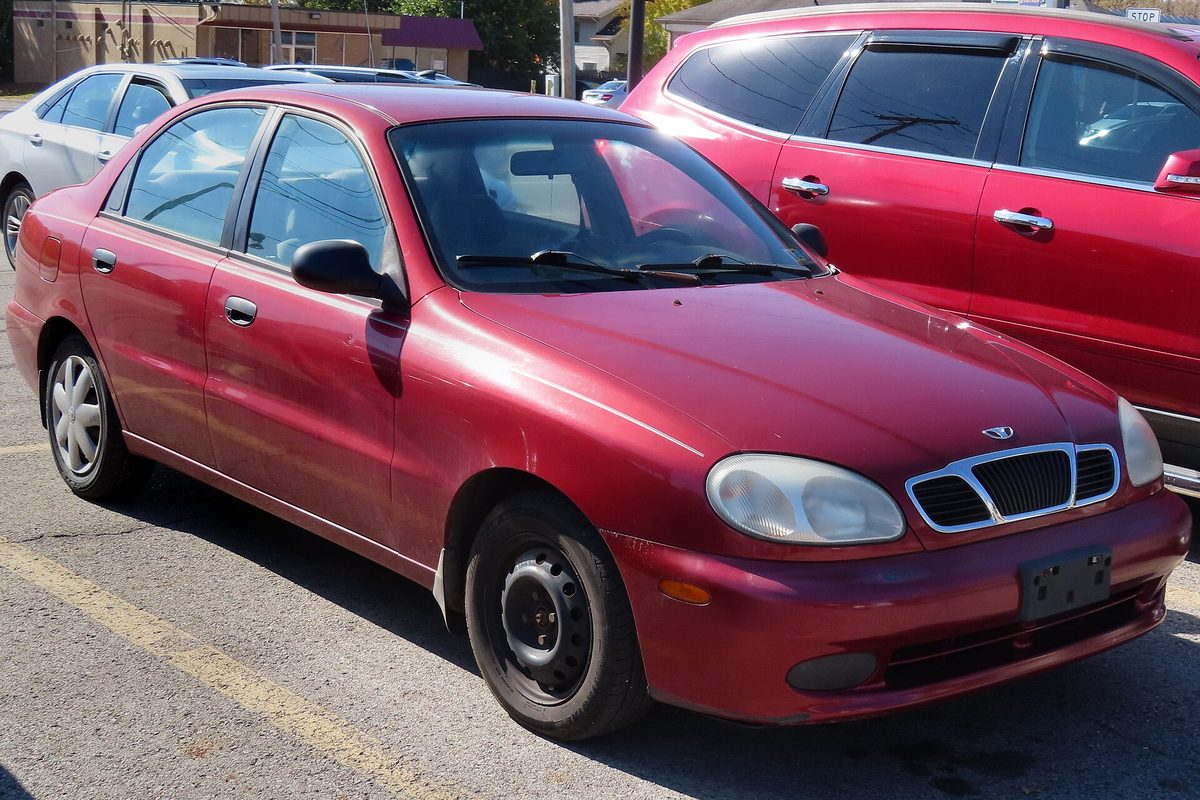 A red Daewoo Lanos sedan on plain steel wheels in a parking lot, next to a red SUV