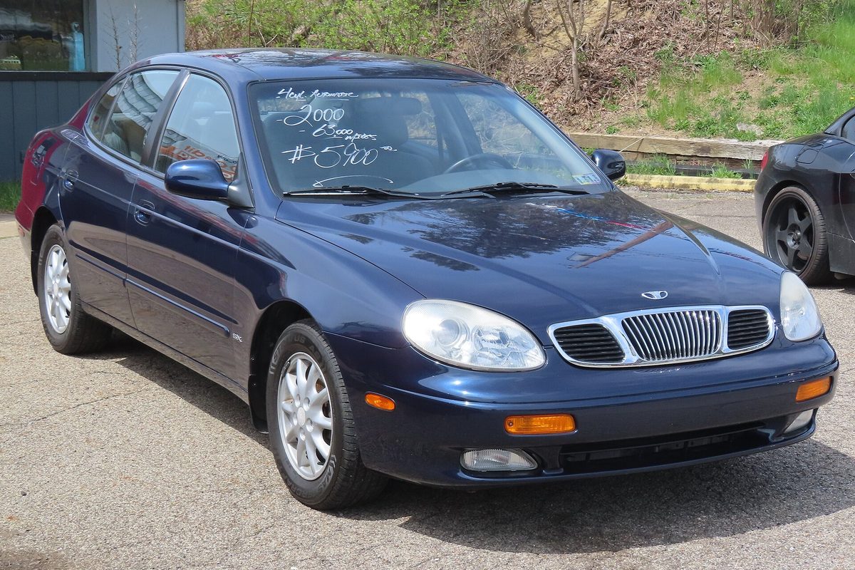 A dark blue Daewoo Leganza sedan on a gravel lot, with white sale writing on its windshield