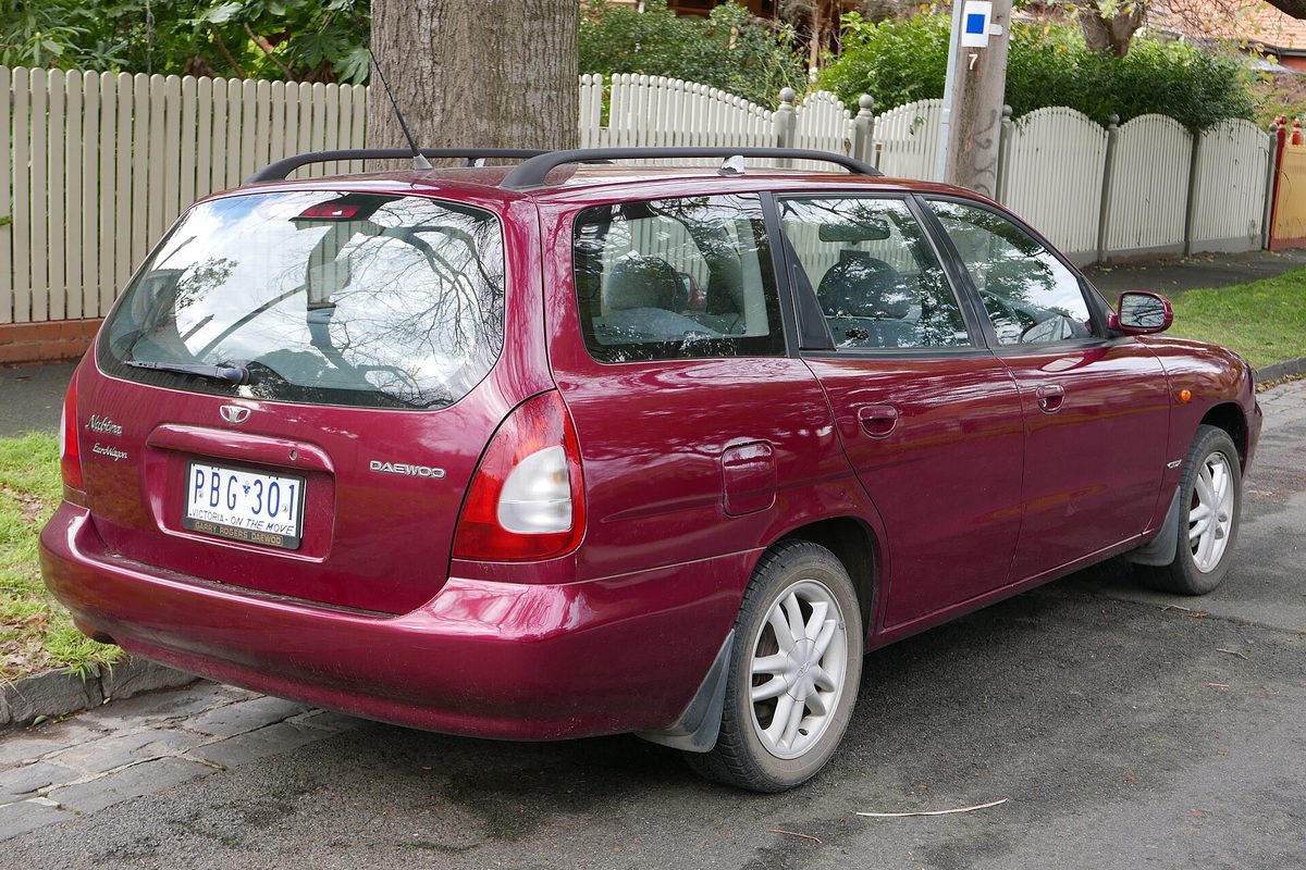 Rear view of a maroon Daewoo Nubira station wagon parked at a curb beside a white picket fence and a large tree trunk