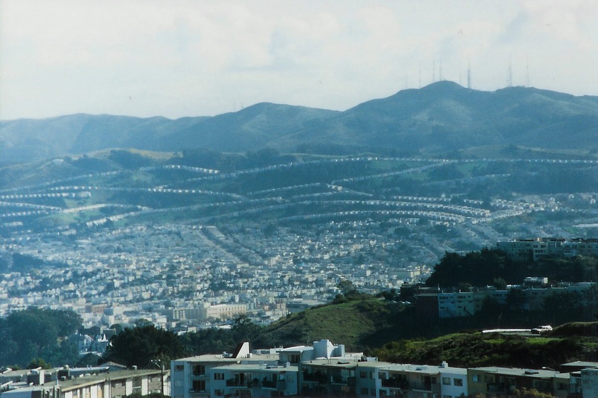 Rows of Daly City houses curving across green hills, with San Bruno Mountain and its antenna towers behind and apartment blocks in front