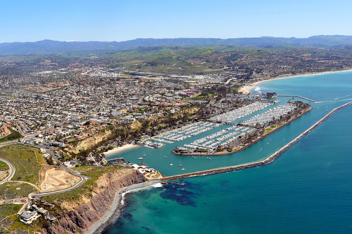 Aerial view of Dana Point Harbor, its marinas packed with boats behind a long rock breakwater, with coastal bluffs, neighborhoods and mountains beyond