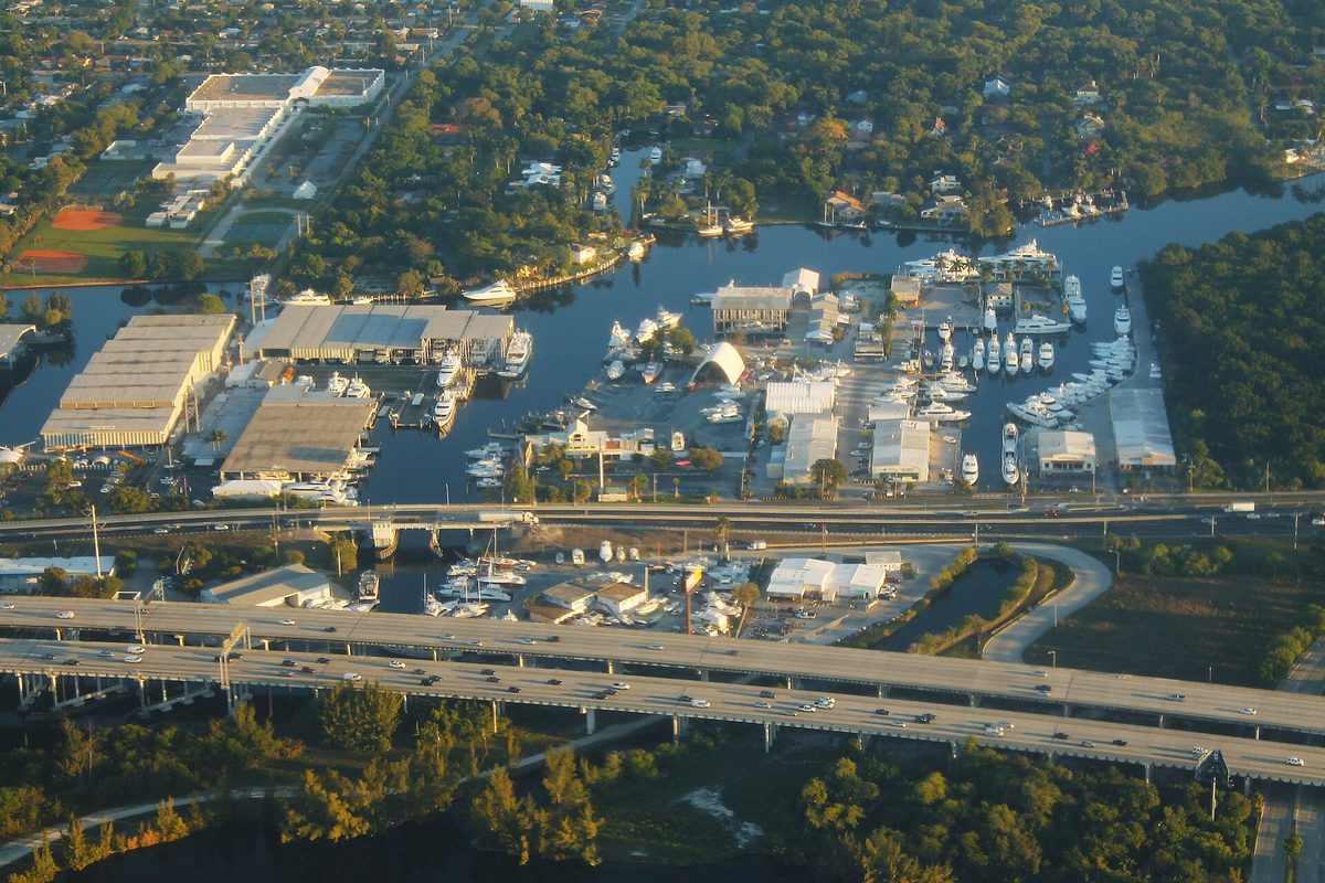 Aerial view of Interstate 95 crossing a river lined with marinas, boatyards and warehouse buildings, with trees and homes beyond