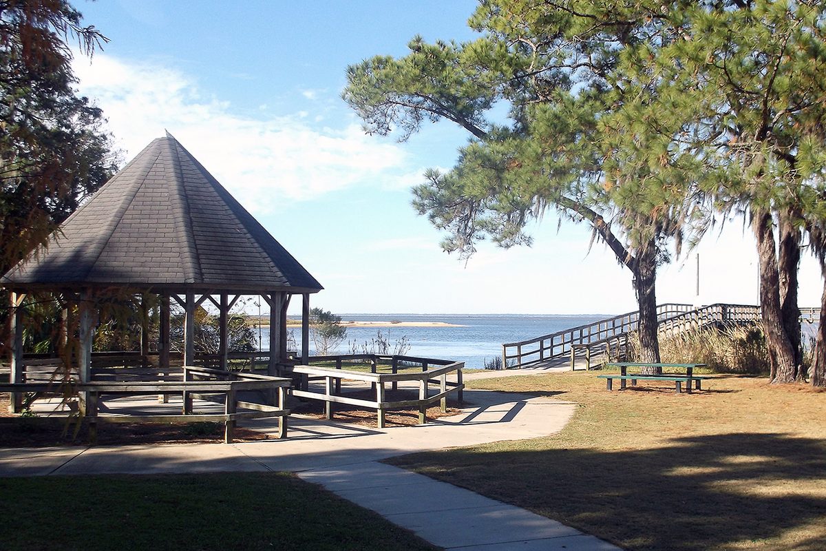 A shingled wooden gazebo and a boardwalk under tall pines at Bayfront Park in Daphne, with Mobile Bay stretching out behind them