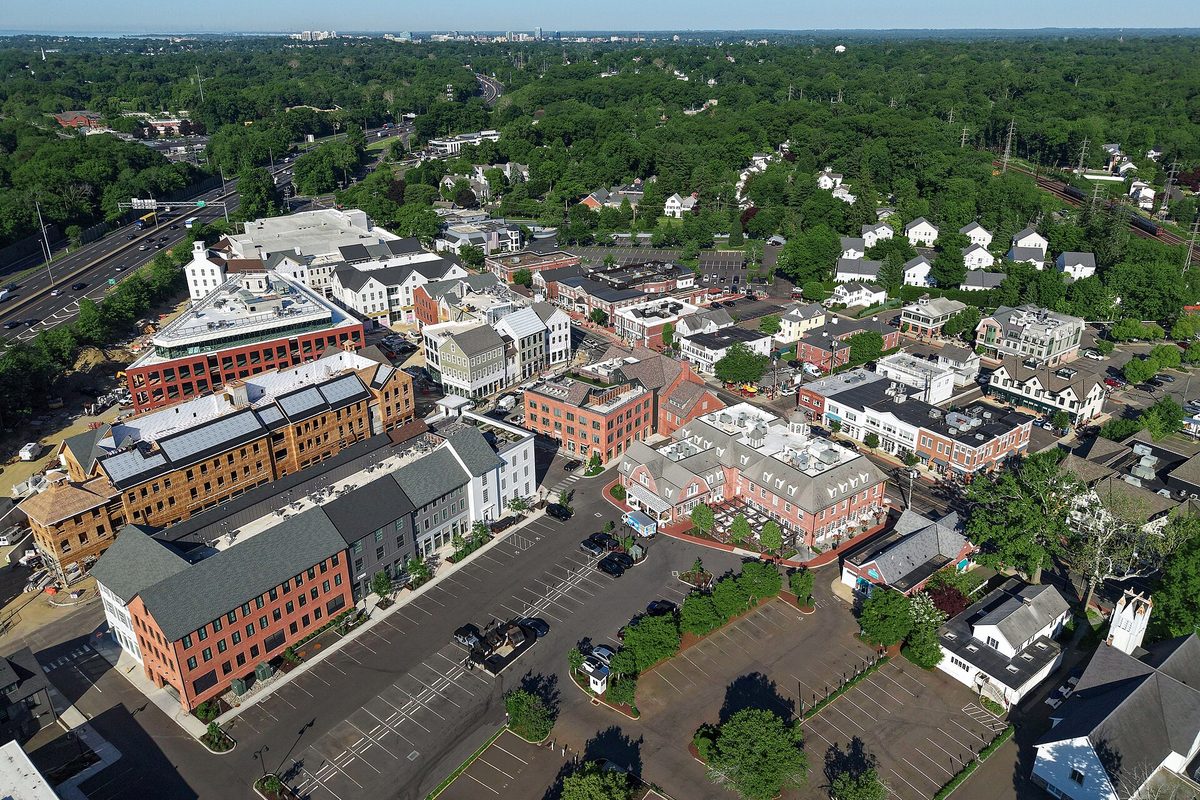 Aerial view of downtown Darien: brick and gray-sided shop and apartment buildings around a large parking lot, a highway on the left and thick trees beyond