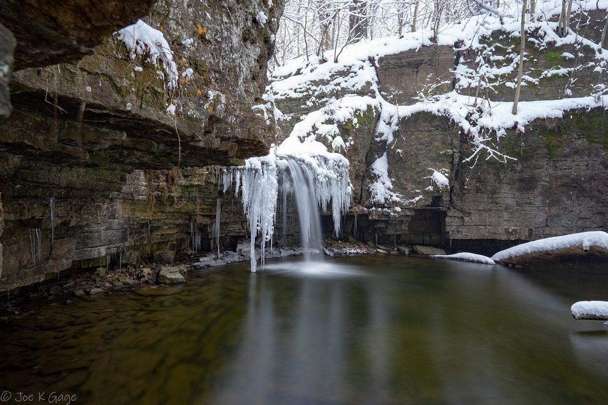 Canyon Creek Falls at Waterfall Glen Forest Preserve in southern DuPage County in winter, a thin waterfall hung with icicles over a rock ledge, with snow on the cliffs