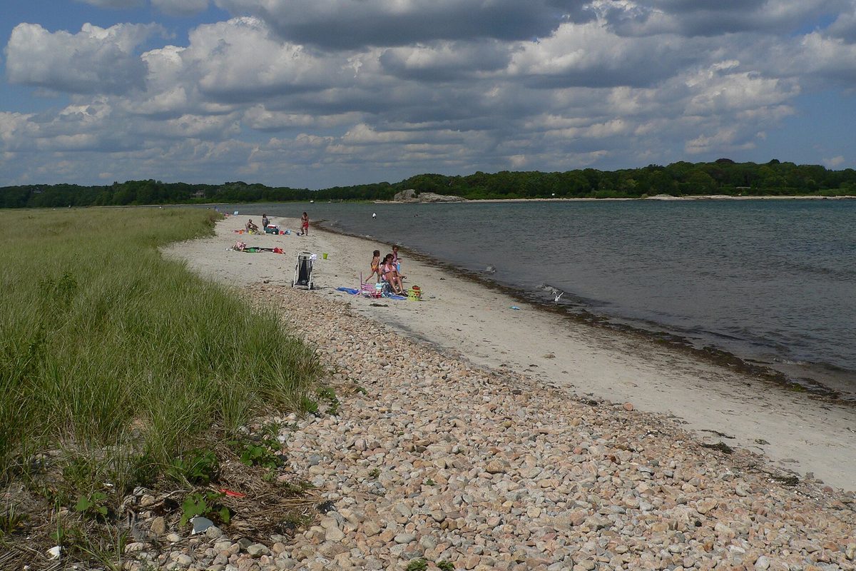 A sand and pebble beach at Demarest Lloyd State Park in Dartmouth, with a few beachgoers near the water and a wooded shore across the bay