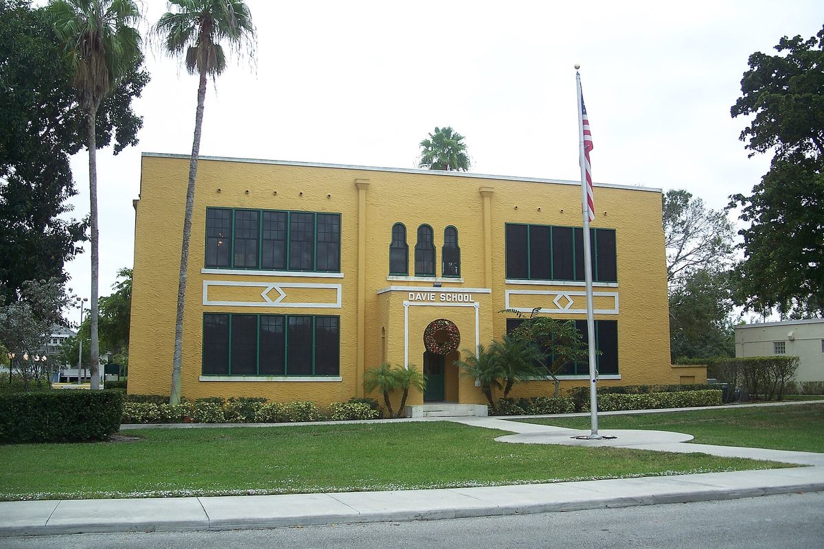 The two-story yellow Davie School building with tall dark windows and an arched entrance, palm trees and a flagpole on the front lawn