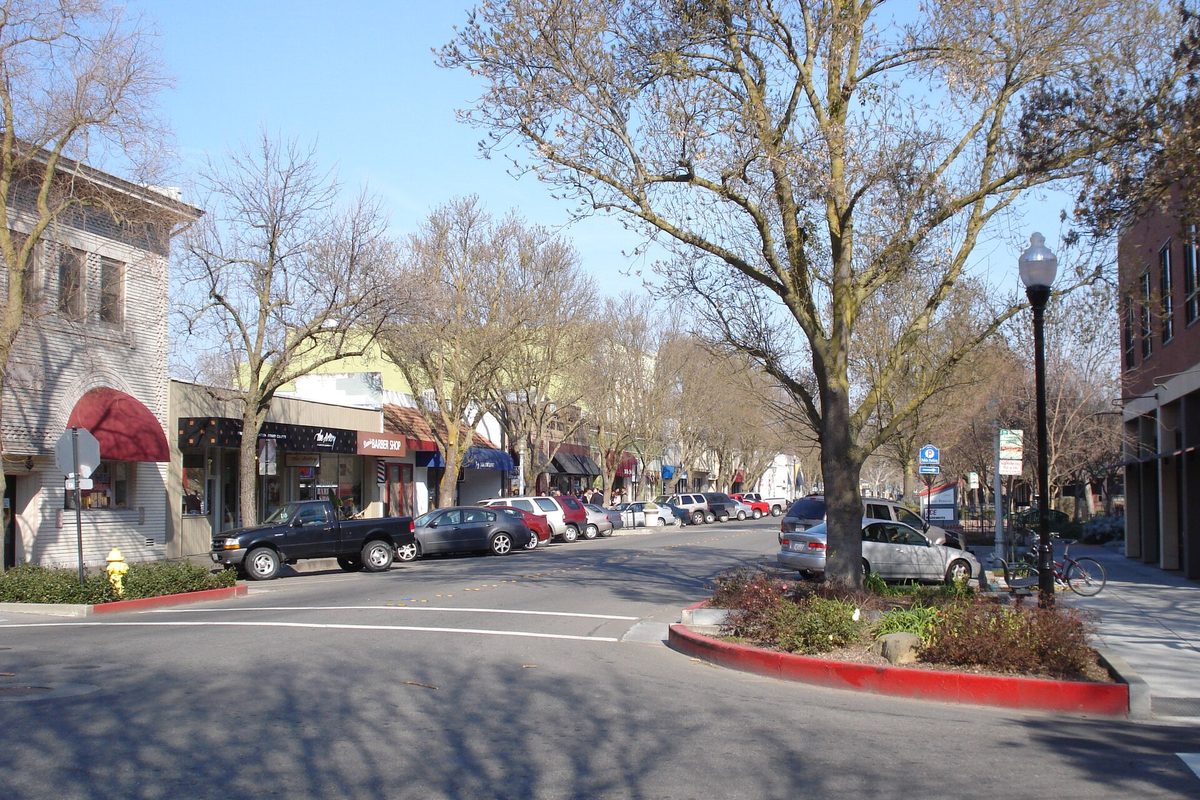 G Street in downtown Davis on a sunny morning, with bare trees, parked cars, storefronts and a red-curbed planter in the foreground