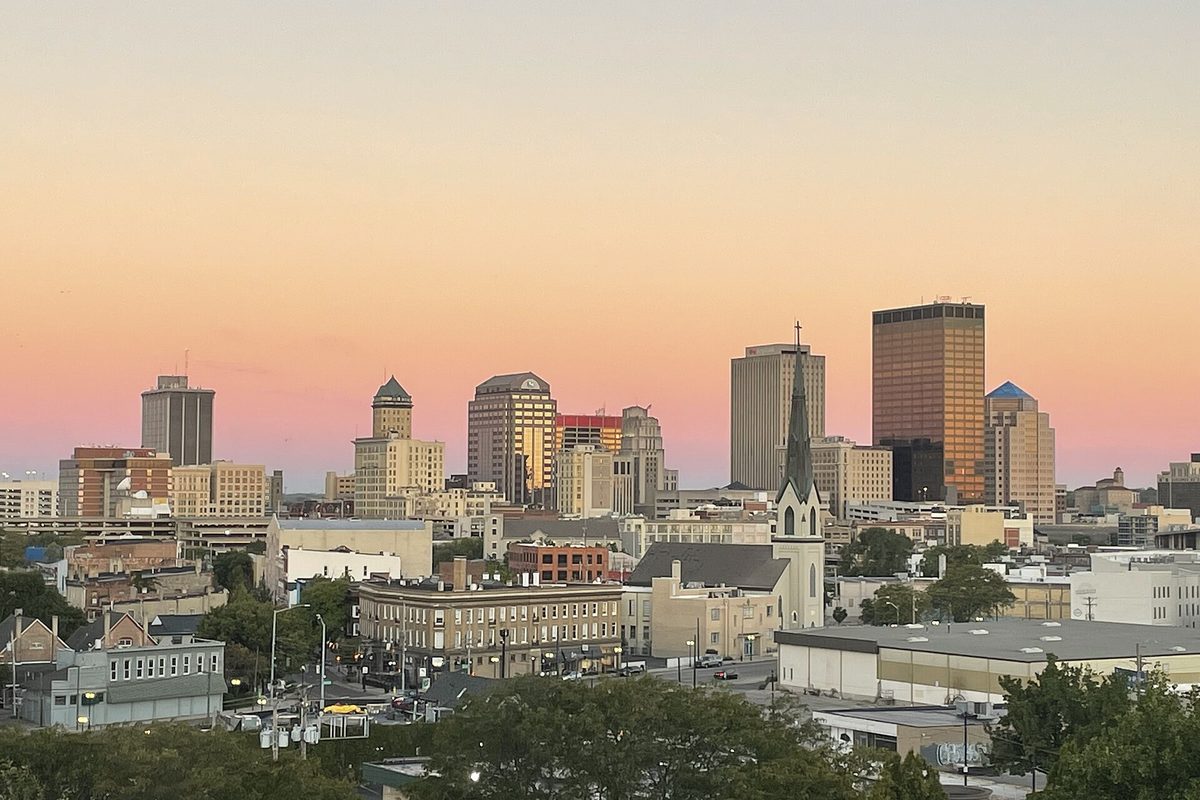 Downtown Dayton's office towers and a church steeple against a pink and orange sunset sky, seen from a high-rise east of downtown