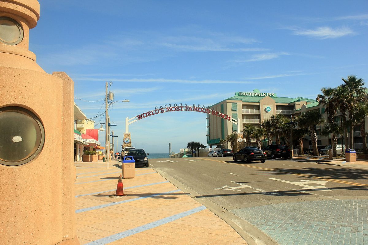 The arch reading World's Most Famous Beach over the road down to the sand in Daytona Beach, with palm trees, a hotel and parked cars beside it