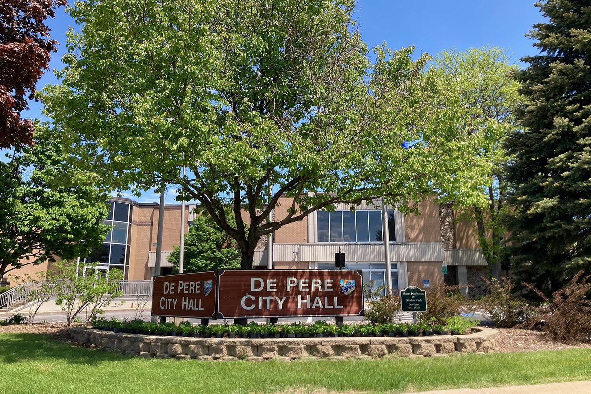 De Pere City Hall, a tan brick building partly hidden behind a large leafy tree, with a brown De Pere City Hall sign in front