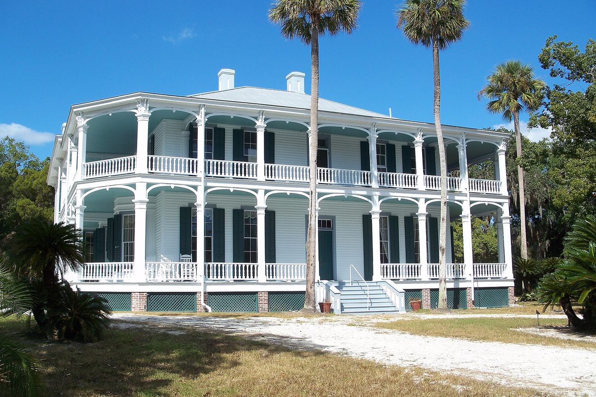 DeBary Hall, a white two-story mansion with wraparound porches on both floors, dark green shutters and tall palm trees beside a sandy drive