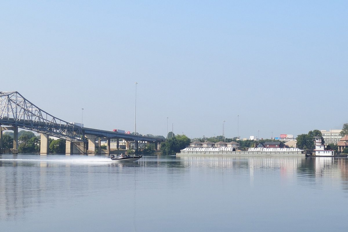 A bass boat speeding across the Tennessee River at Decatur past a towboat with covered barges, a steel truss bridge carrying traffic on the left