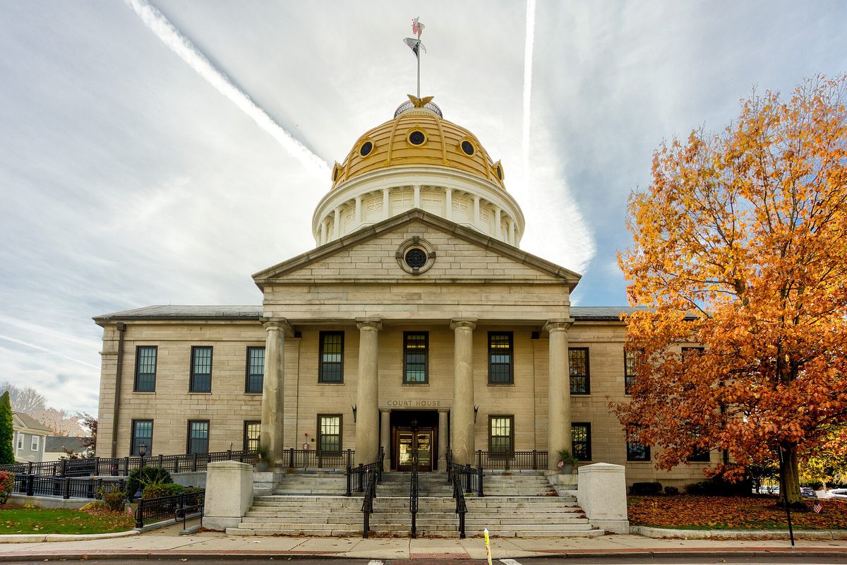 The granite Norfolk County Courthouse in Dedham, with stone columns and a gold dome, beside a tree in orange autumn leaves