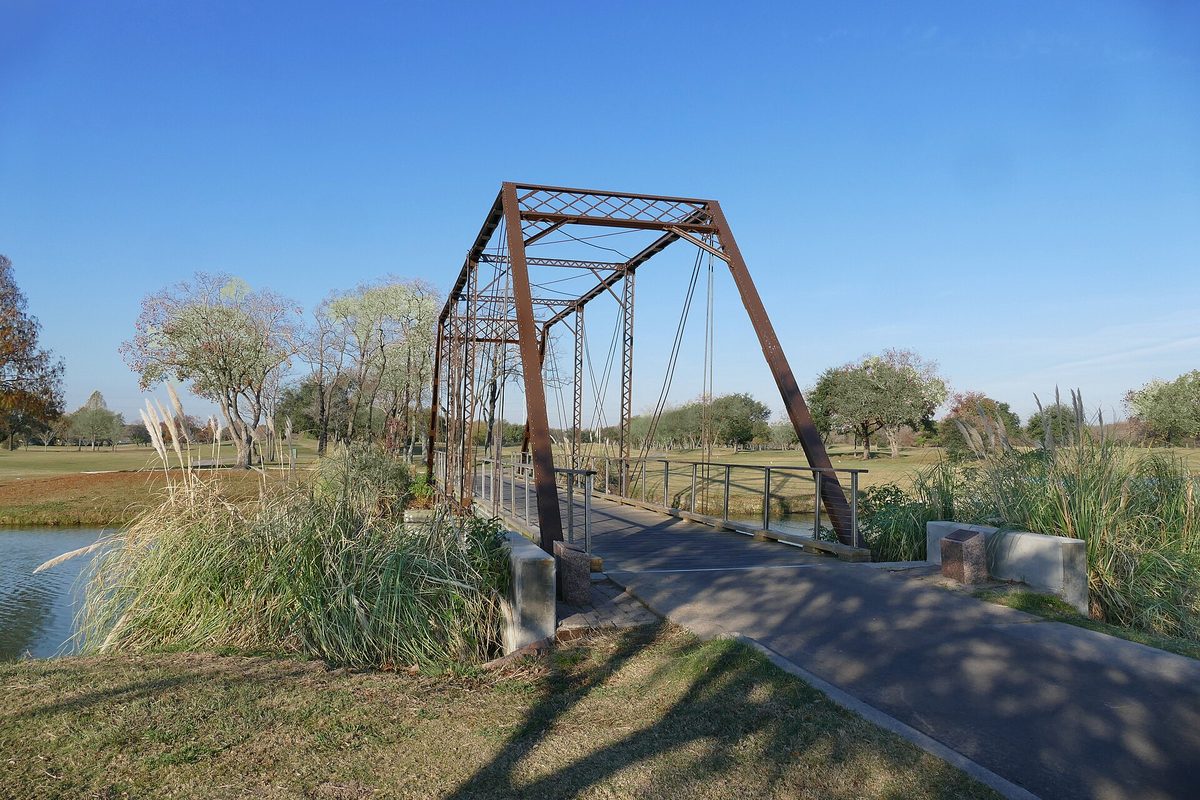A rust-brown iron truss footbridge over a pond in Deer Park, with tall pampas grass and trees under a clear blue sky