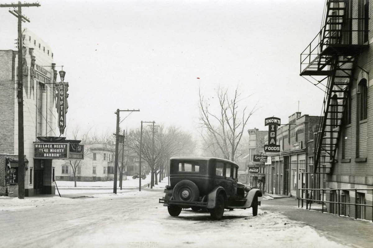Old black-and-white winter photo of a snowy DeKalb street, with a 1930s sedan in the road, the Egyptian Theatre marquee on the left and a Snow's IGA Foods sign on the right