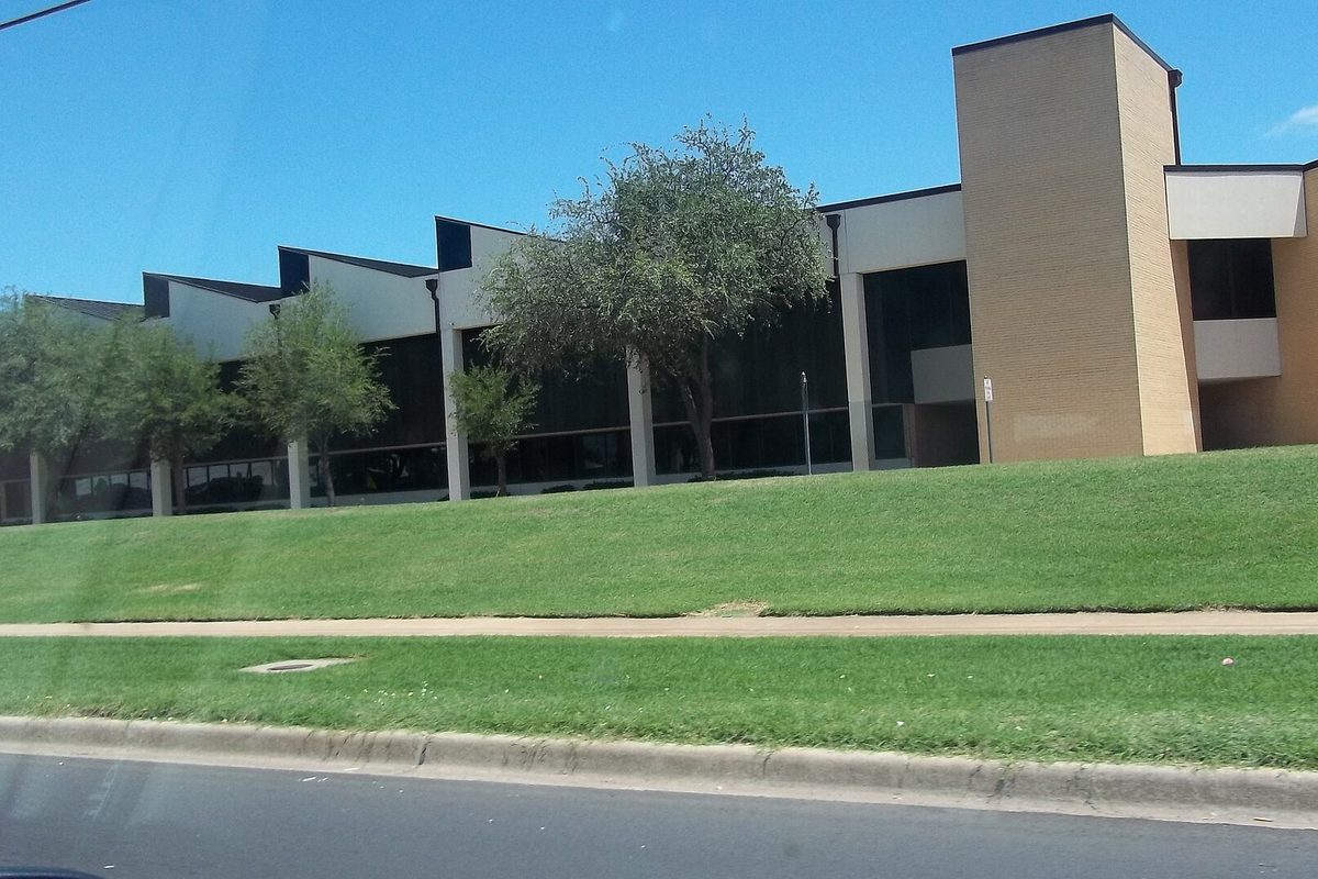 The tan brick and dark glass front of Del City High School behind a sloping green lawn and sidewalk, seen from the street
