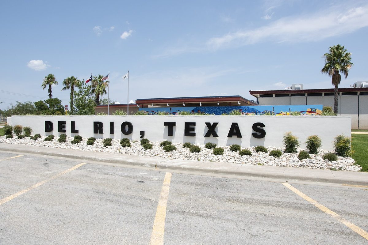 A long white wall with black letters reading Del Rio, Texas, edged with white rocks and shrubs, with palm trees, flags and the civic center behind it