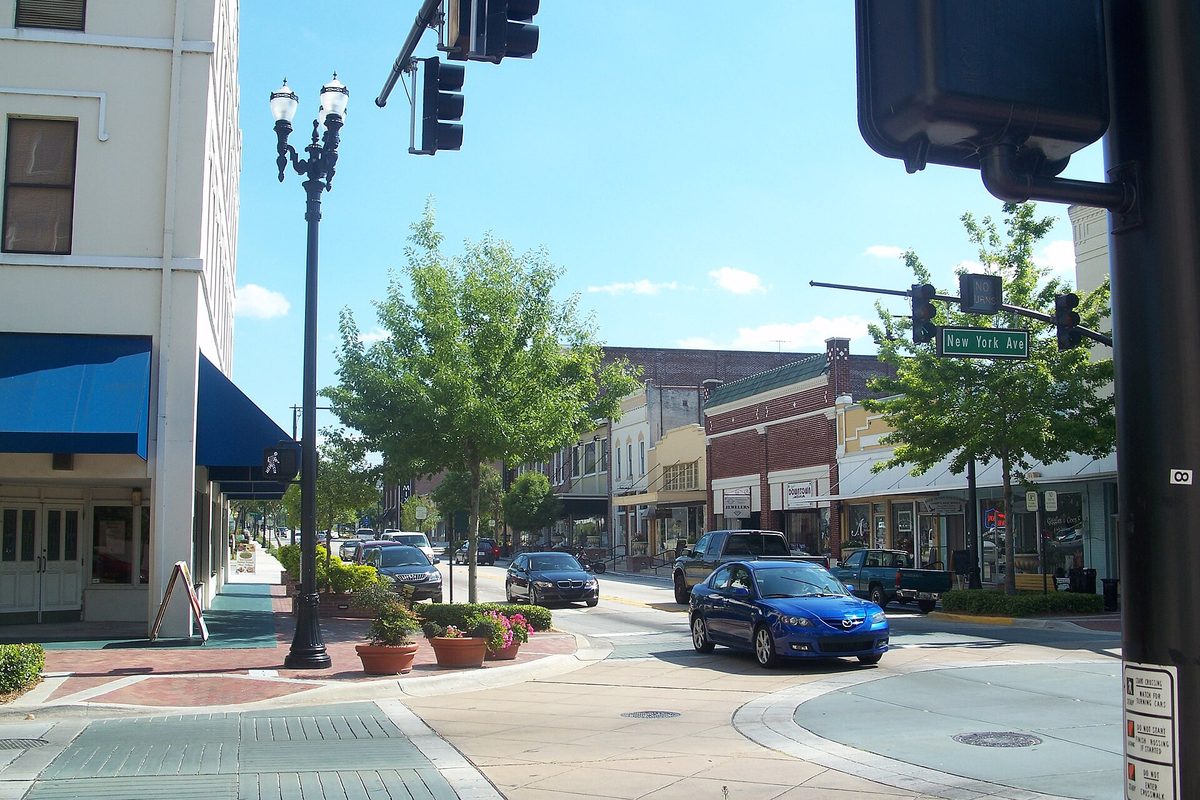Downtown DeLand looking south along a main street of brick storefronts, with a blue hatchback at the corner of New York Avenue and old-style street lamps