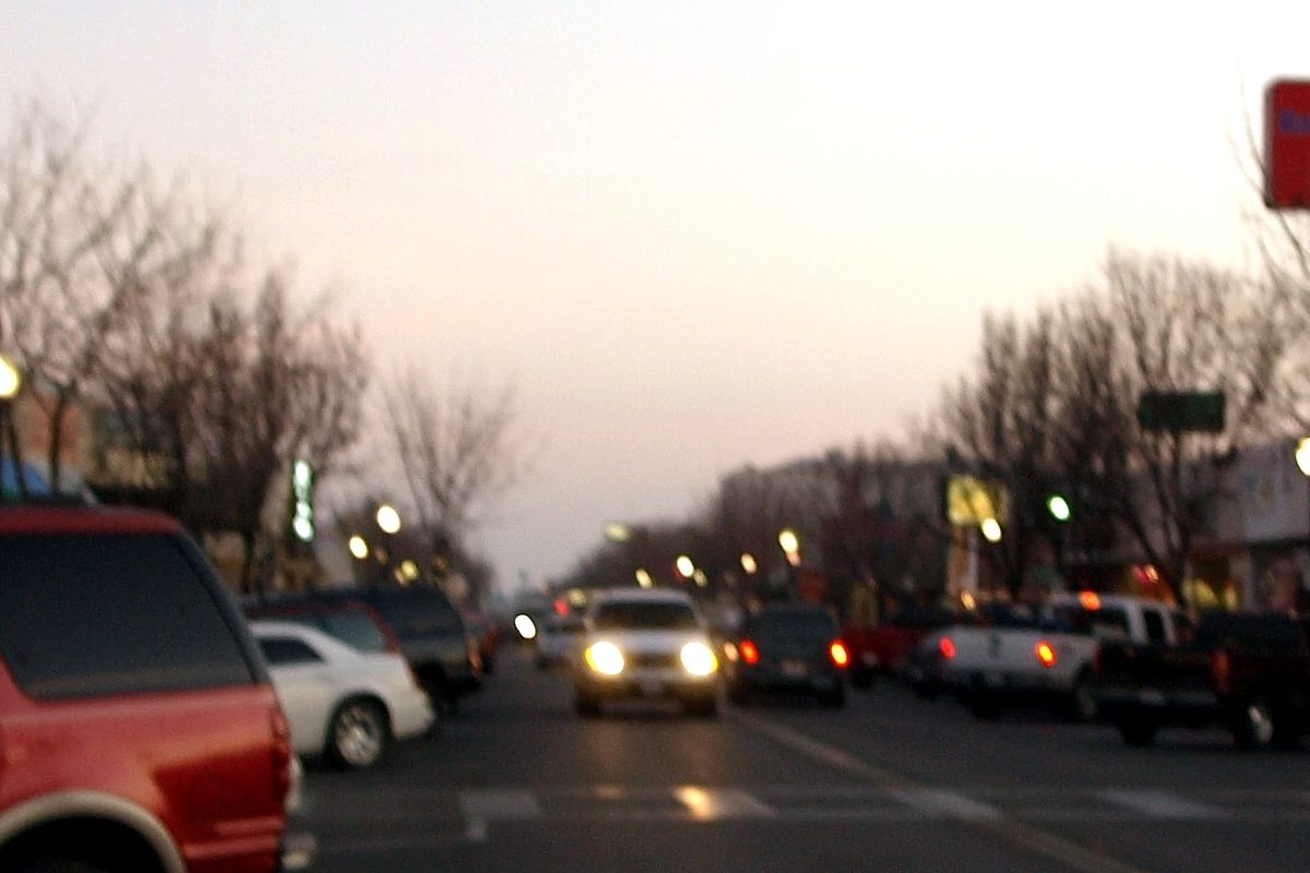 Cars and pickups with headlights on along Main Street in downtown Delano at dusk, bare trees lining the street