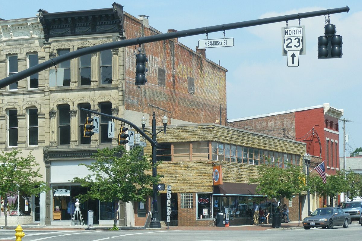 Brick and stone storefronts at a downtown Delaware, Ohio intersection, under traffic lights hung with signs for North Sandusky Street and U.S. Route 23 north