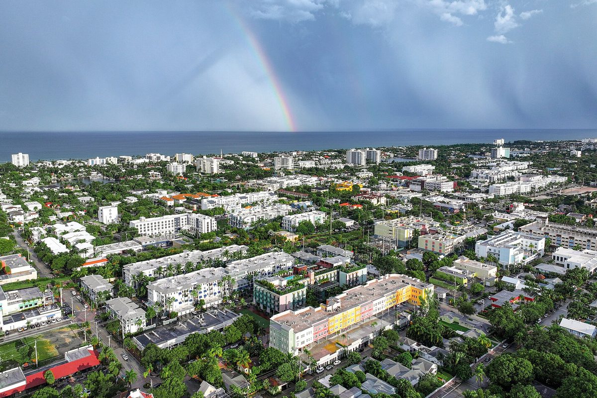 Aerial view over Delray Beach toward the Atlantic Ocean, with a rainbow above low-rise buildings, tree-lined streets and a colorful building in front