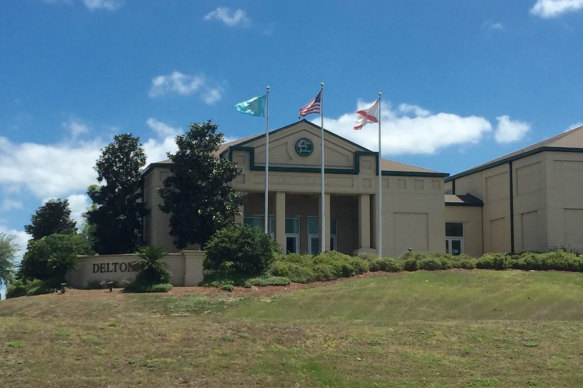 Deltona City Hall, a tan building with a columned entrance and three flags flying, on a grassy rise beside a Deltona sign