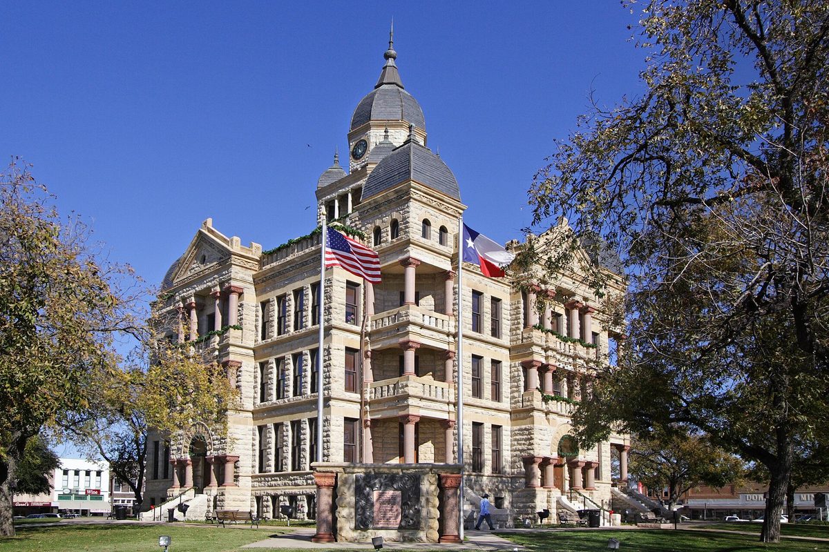 The Romanesque Denton County Courthouse, a pale stone building with a domed clock tower, flying the U.S. and Texas flags under a clear sky
