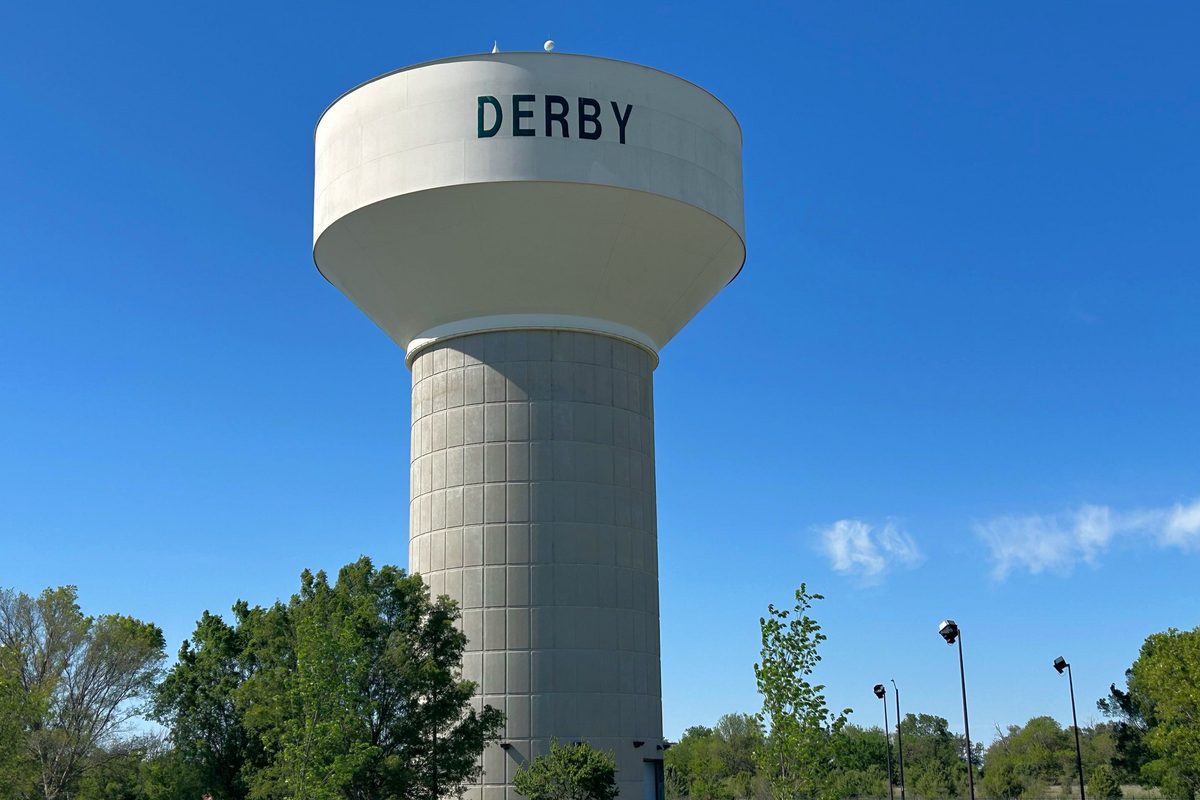 A tall concrete water tower with DERBY painted across the tank, against a clear blue sky above green trees and lamp posts