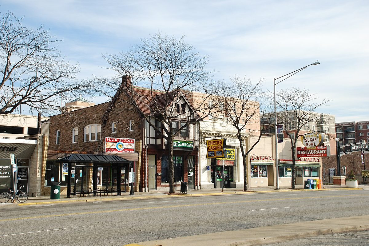 A row of older brick and Tudor-style storefronts on a Des Plaines street, including a hair studio, a currency exchange and the Sugar Bowl restaurant, with bare trees and a bus shelter