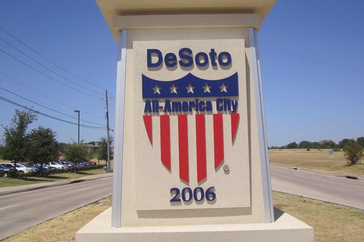DeSoto All-America City 2006 sign with a red, white and blue shield on a cream pillar beside a divided road under a clear sky