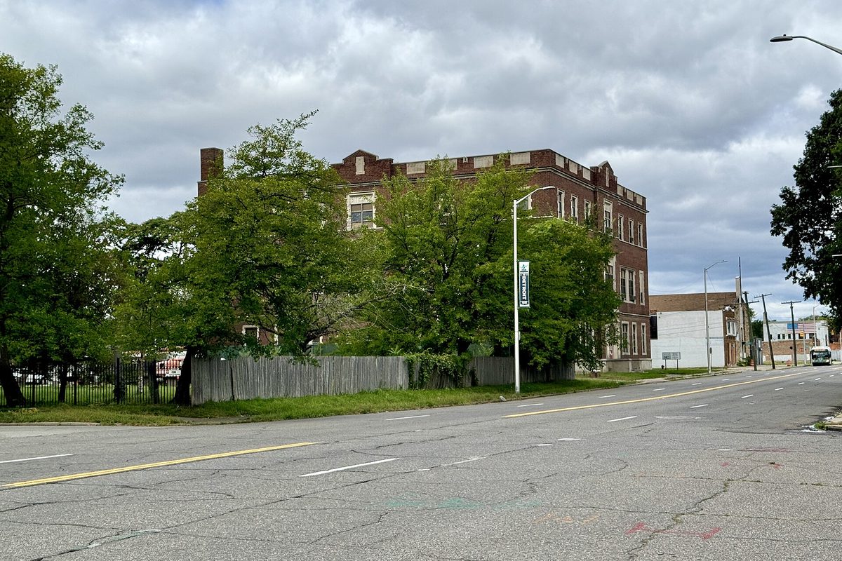 A three-story brick building behind trees and a weathered wooden fence along a wide, cracked street on Hamilton Avenue in Detroit, under a cloudy sky