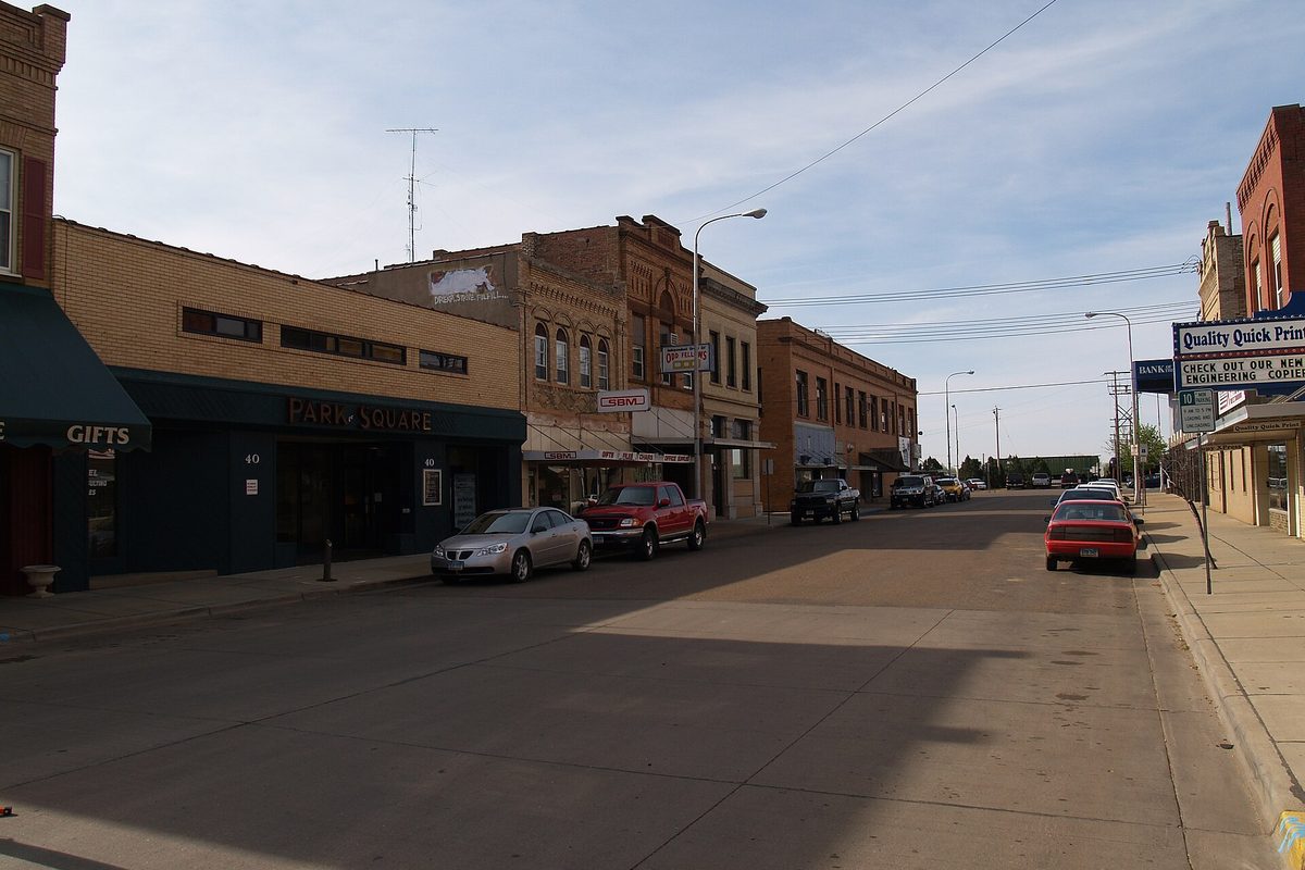A downtown Dickinson street lined with old brick storefronts, with a silver sedan, a red pickup and other cars parked along the curb