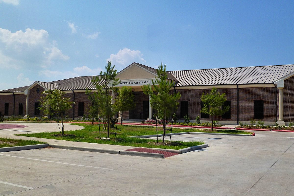 Dickinson City Hall, a long brick building with a metal roof and a white columned entrance, behind young trees and an empty parking lot