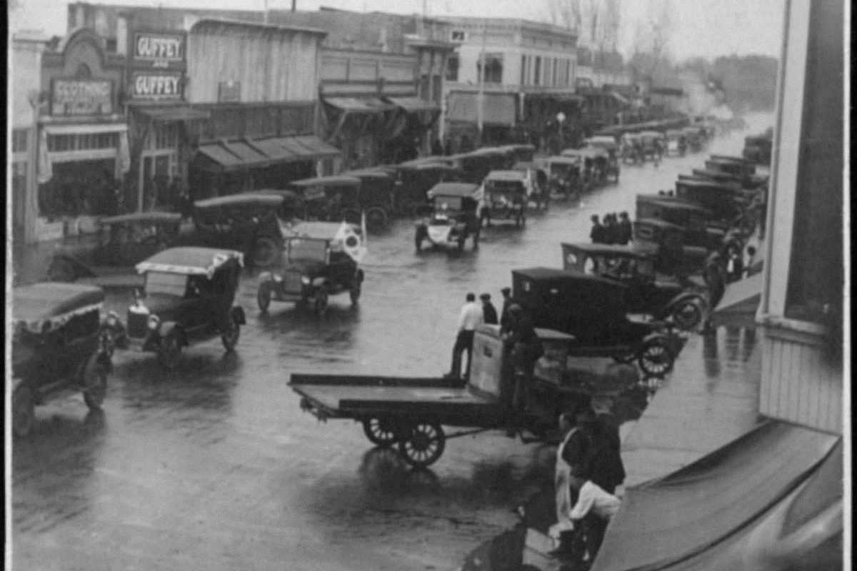 Black-and-white 1920 photo of a parade in Dinuba, with early automobiles parked along a wet main street, storefronts behind them and a flatbed cart in the foreground
