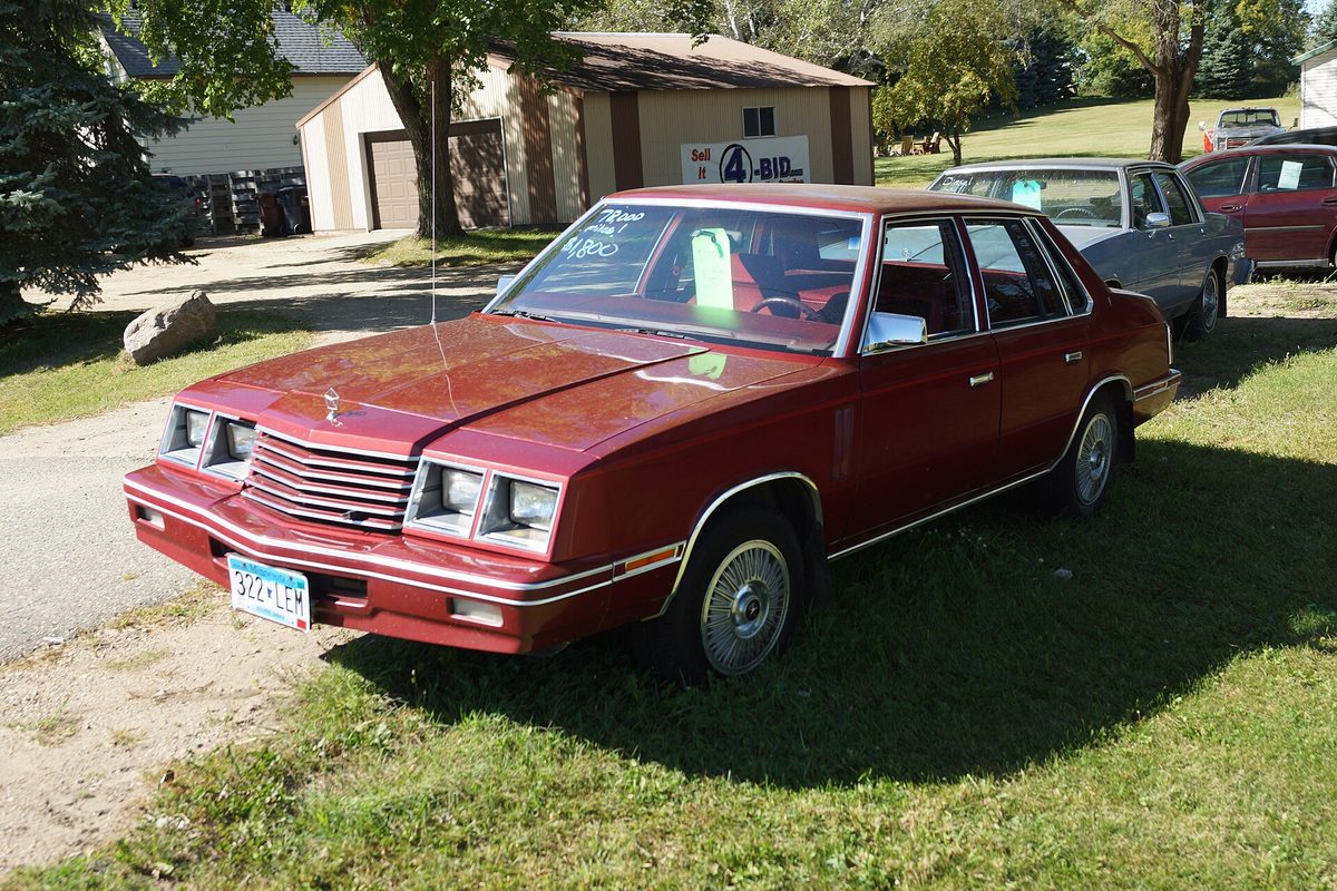 A maroon Dodge 600 four-door sedan parked on grass with handwritten notes inside the windshield