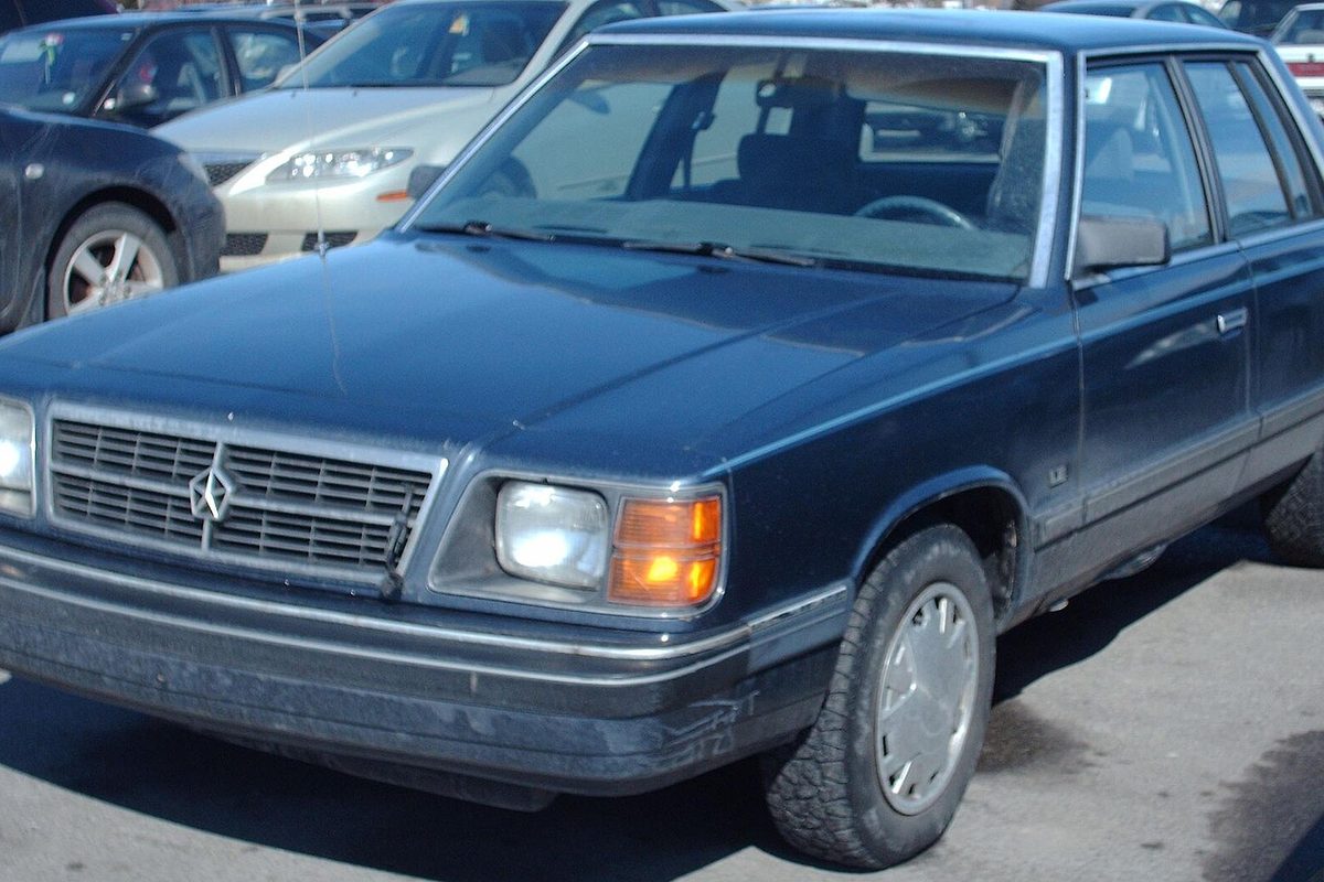 A dark blue Dodge Aries four-door sedan with a chrome grille and amber corner lights, parked in a lot beside other cars
