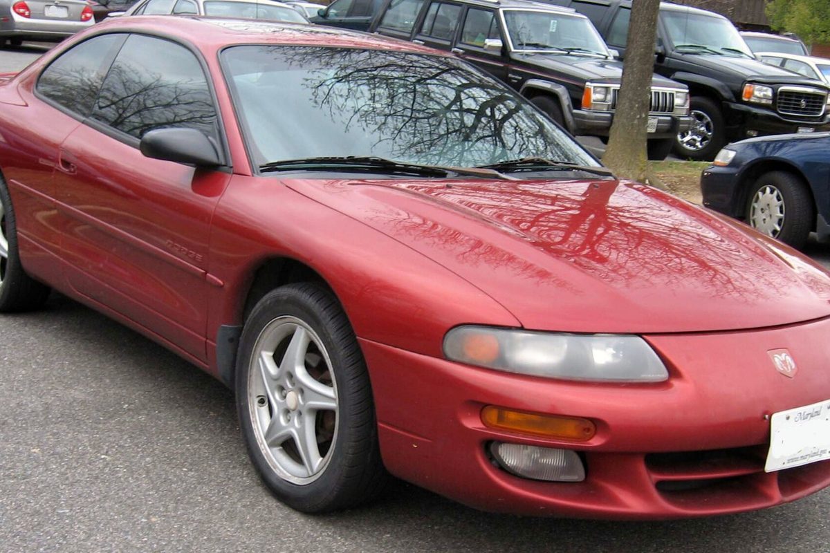 A red 1995-2000 Dodge Avenger two-door coupe with silver alloy wheels in a parking lot, bare branches reflected in the windshield