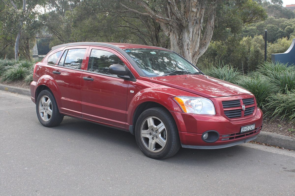 A red 2008 Dodge Caliber SX hatchback parked at a curb in front of trees and shrubs