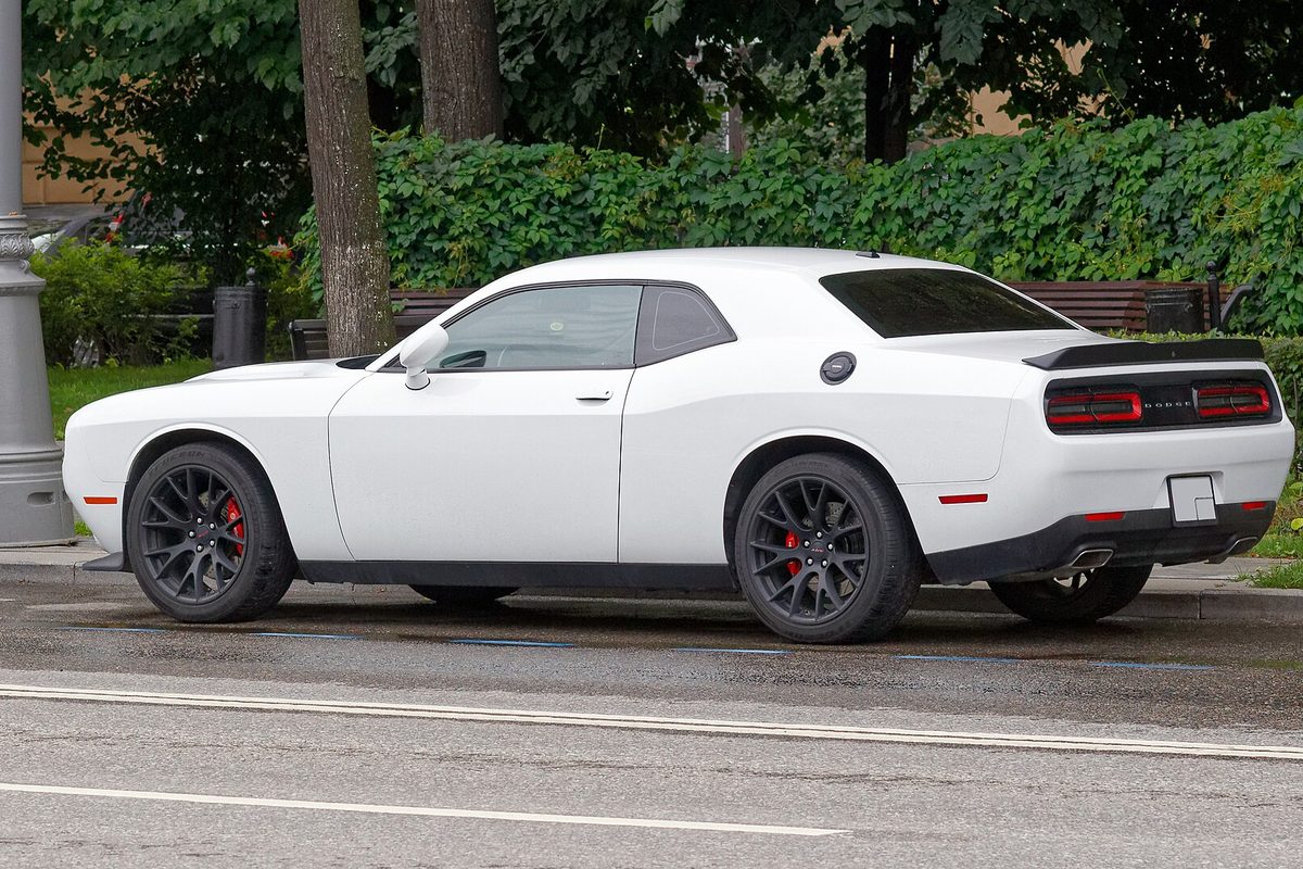 A white Dodge Challenger coupe with dark wheels and red brake calipers, seen from the rear quarter on a wet city street